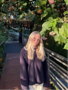 Heather Alden is a student dietitian, a young white woman with blonde hair, posing in sunlight in the garden. She writes about diet culture and politics. 
